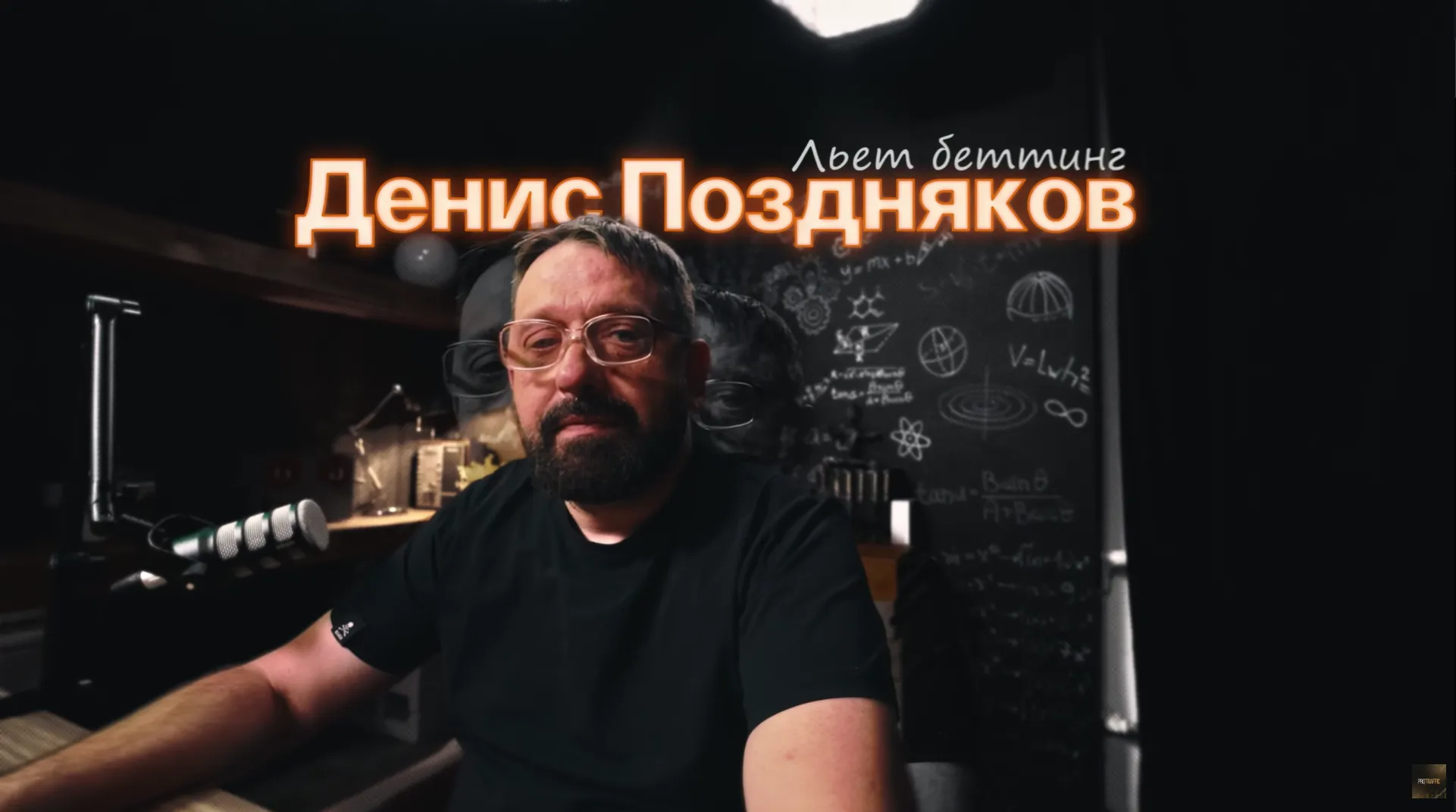 Человек с бородой и очками сидит за столом в студии, окружённый предметами для работы.