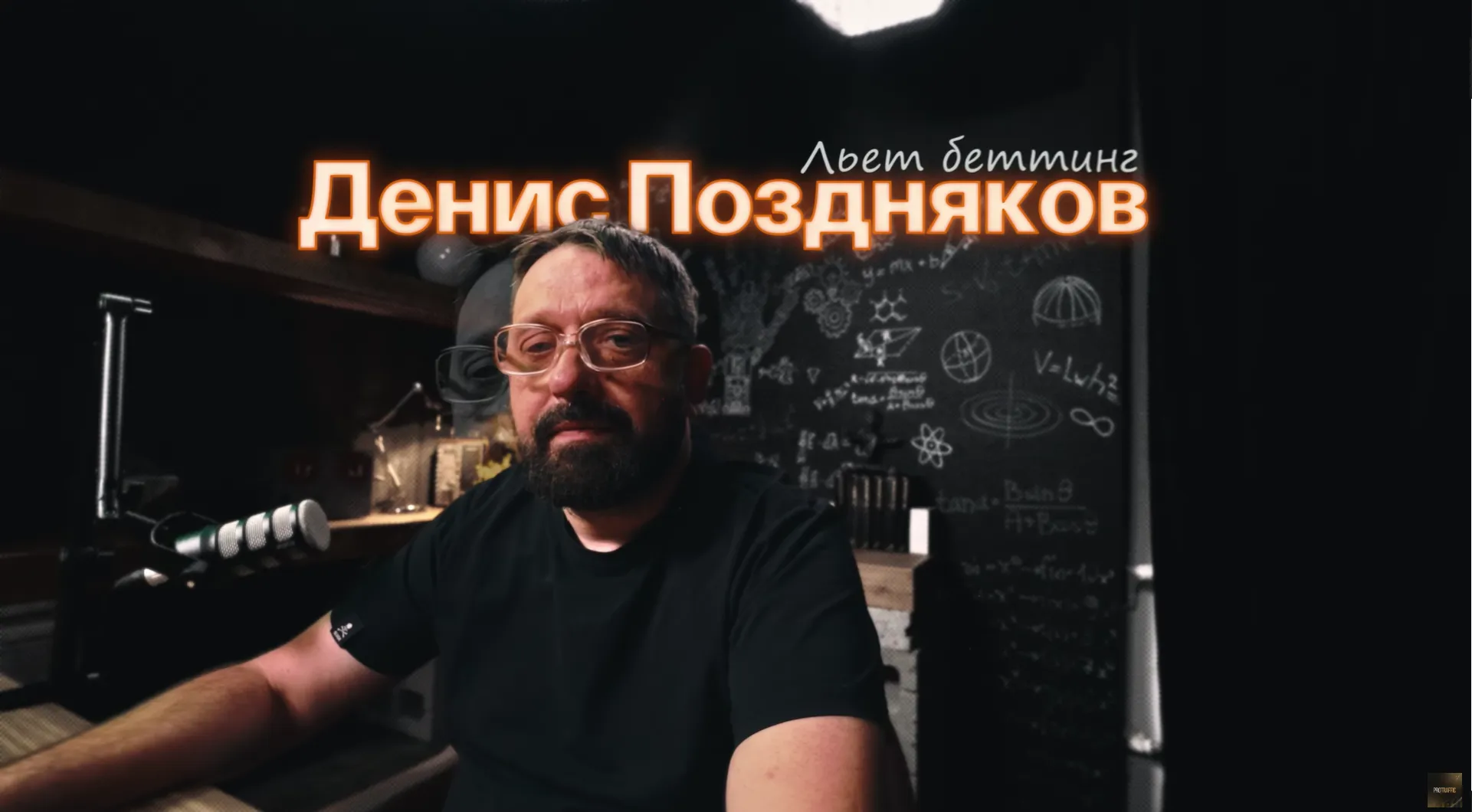 Мужчина с бородой и очками сидит за столом в студии, окружённый предметами для съёмки.